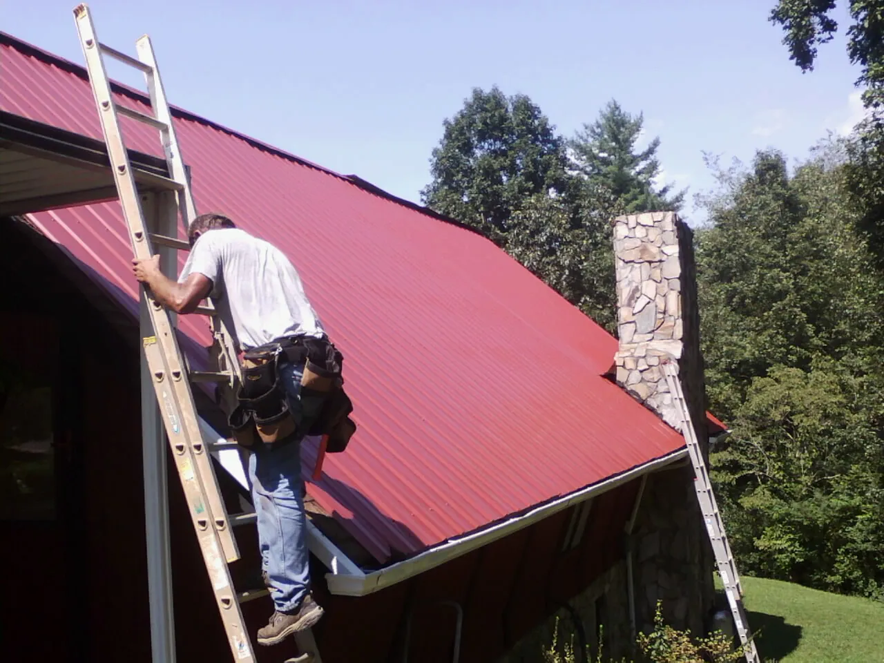 Roofer on a ladder beside a red metal roof and stone chimney — Soffit & Fascia Repair by Roofing Brothers in Canyonville